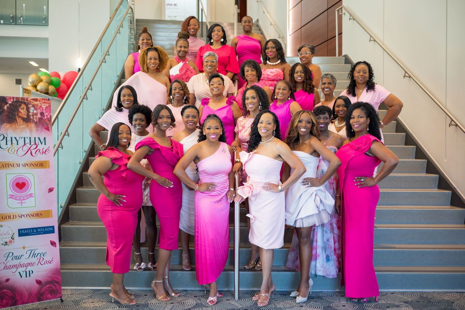 Large group of women in pink and fuchsia dresses pose together on grand staircase at event.