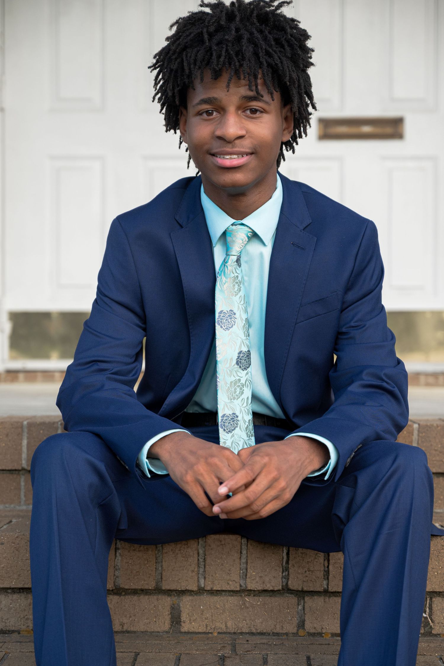 Young man in navy blue suit and teal tie sits smiling on brick steps outside white door.