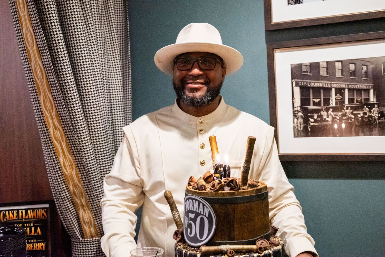 Smiling man in white outfit and hat celebrating 50th birthday holding decorated chocolate cake.