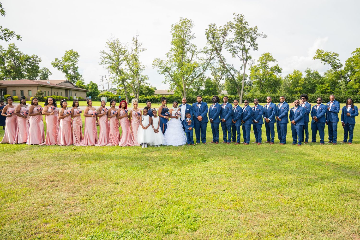 Large wedding party with bridesmaids in pink dresses and groomsmen in navy suits on green lawn.