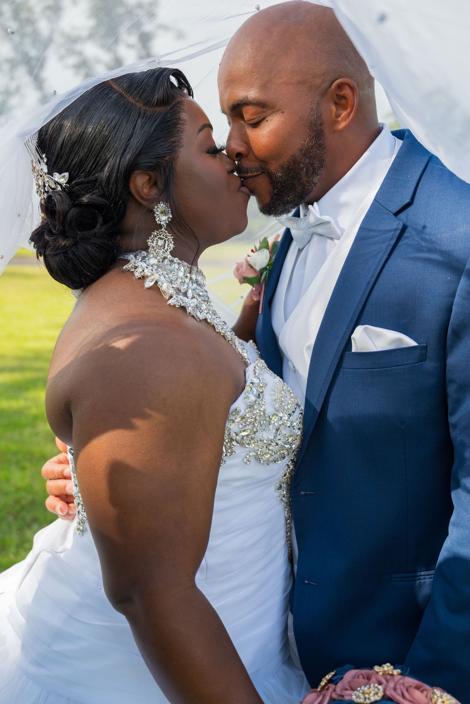 Black bride and groom sharing romantic kiss under veil on sunny wedding day outdoors.