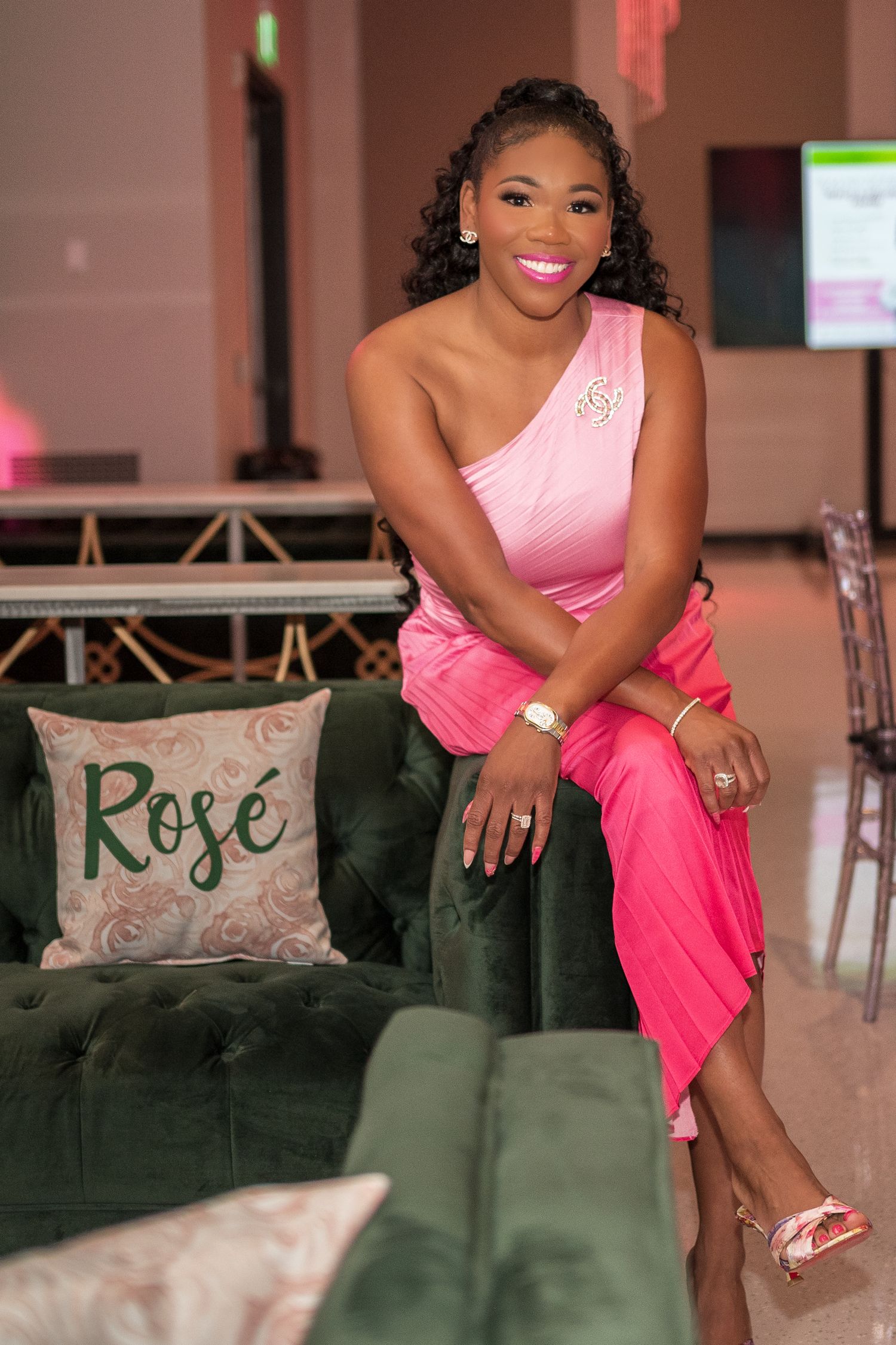 Woman in pink one-shoulder gown sits elegantly on green velvet sofa at formal event.