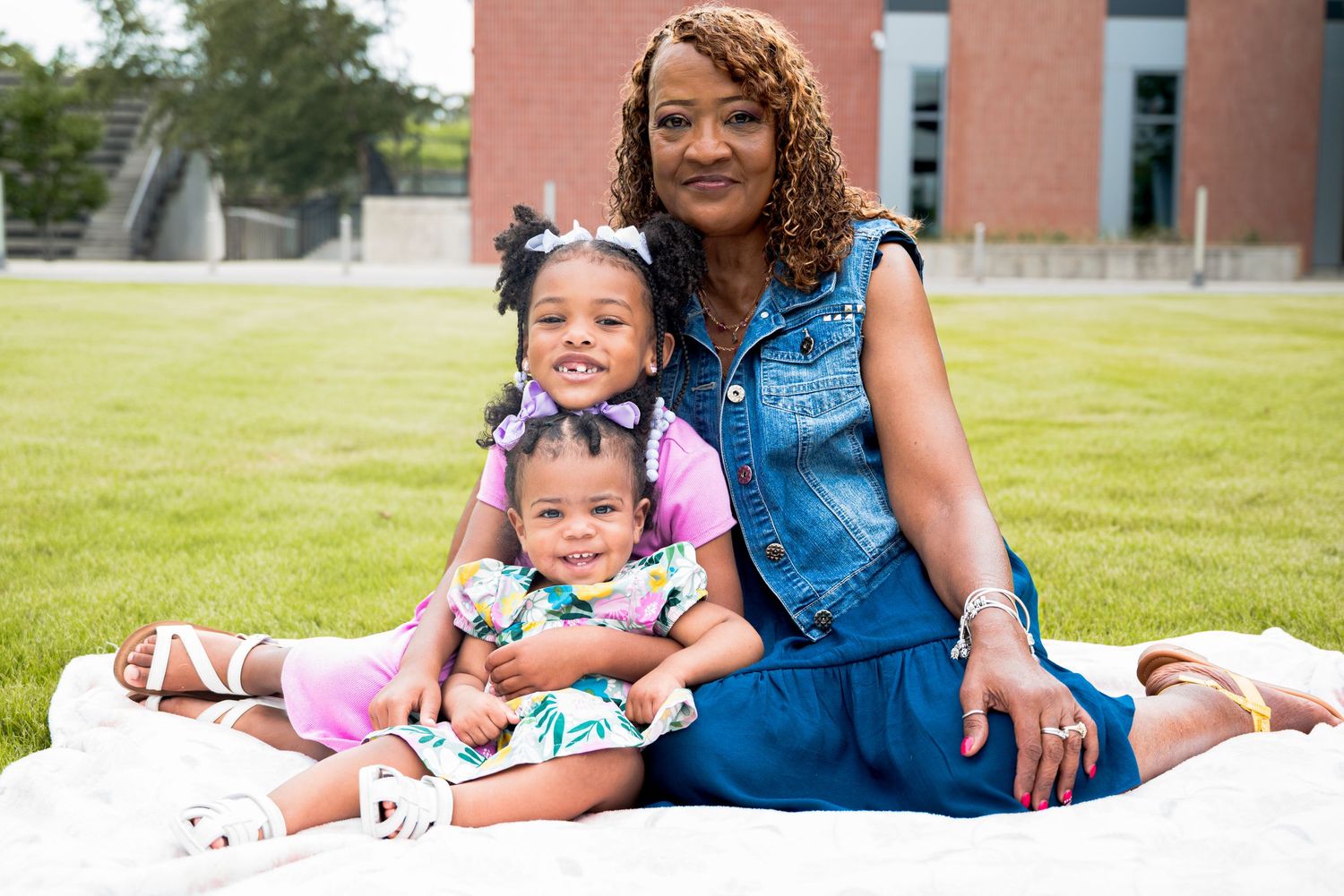 Smiling woman in denim dress sits on white blanket outdoors with two young girls.