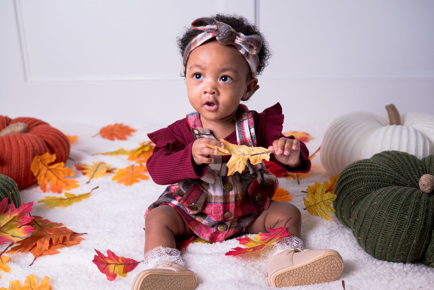 Adorable baby girl in fall plaid dress holding autumn leaf surrounded by colorful pumpkins and leaves.