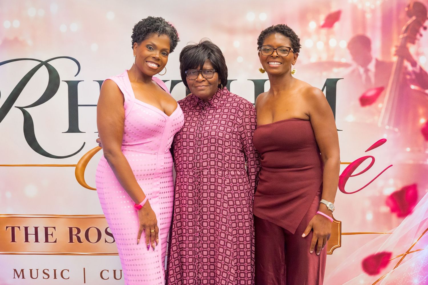 Three women in pink and burgundy dresses pose together at Rhythm and Rose event backdrop.