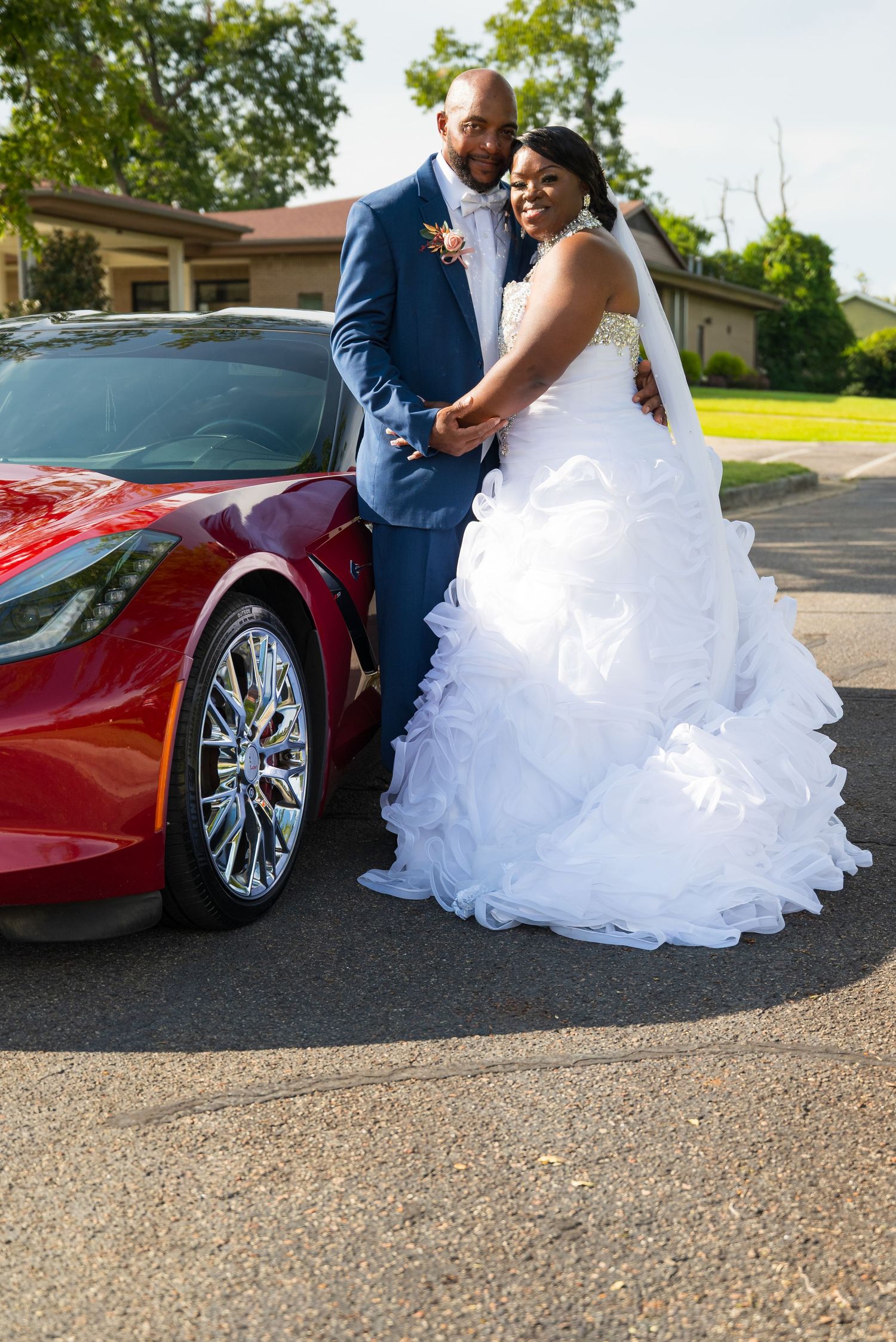 Bride and groom posing elegantly beside a red Corvette sports car on their wedding day.