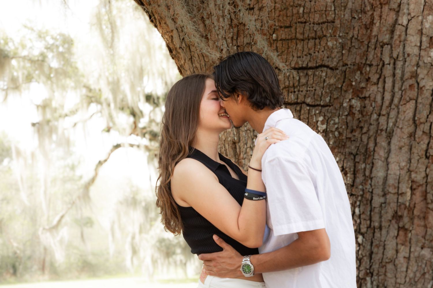 A young couple sharing a romantic kiss against a moss-draped tree in a sunlit outdoor setting.