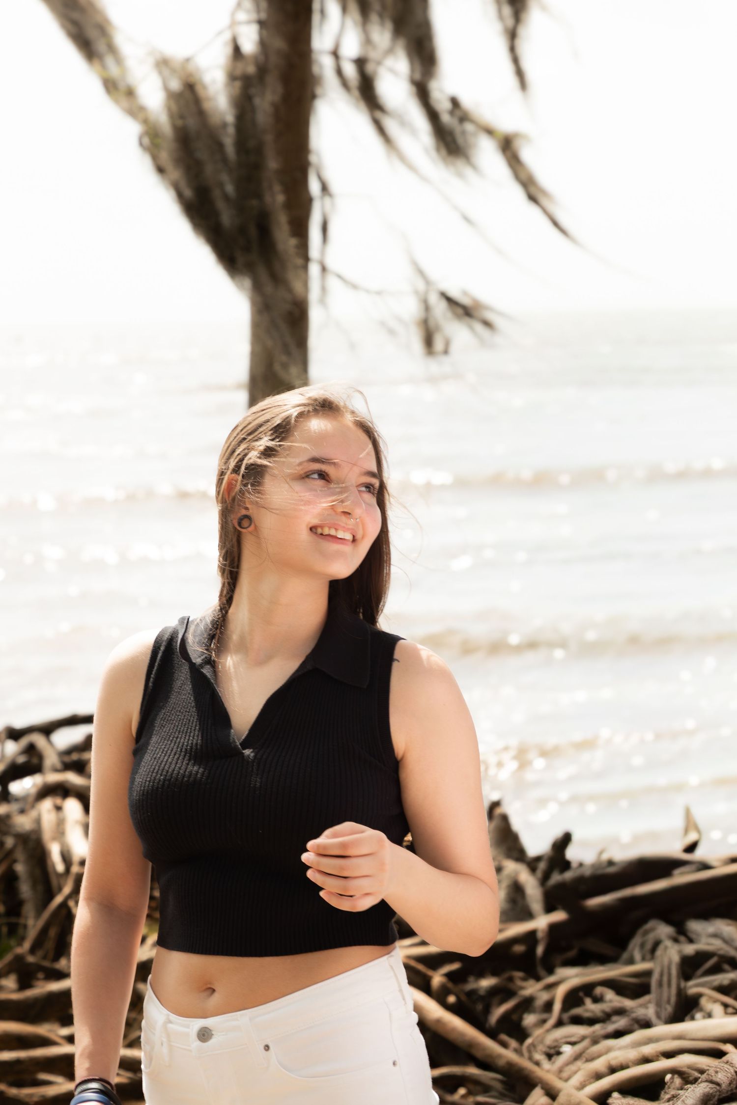 Young woman in black crop top smiling on a sunny beach with driftwood and ocean in the background.