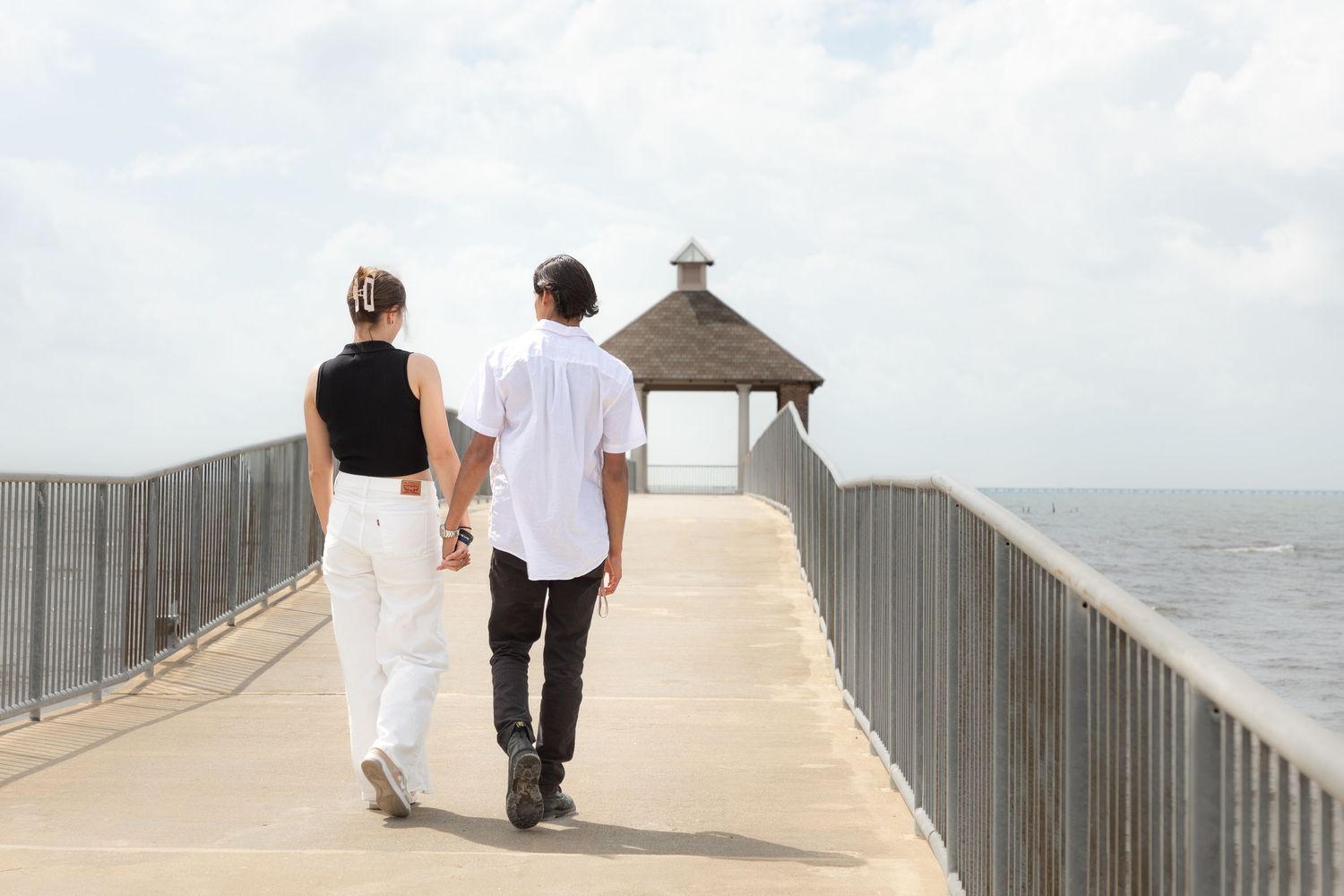 Couple holding hands walking along a scenic pier with a gazebo and ocean views on a sunny day.