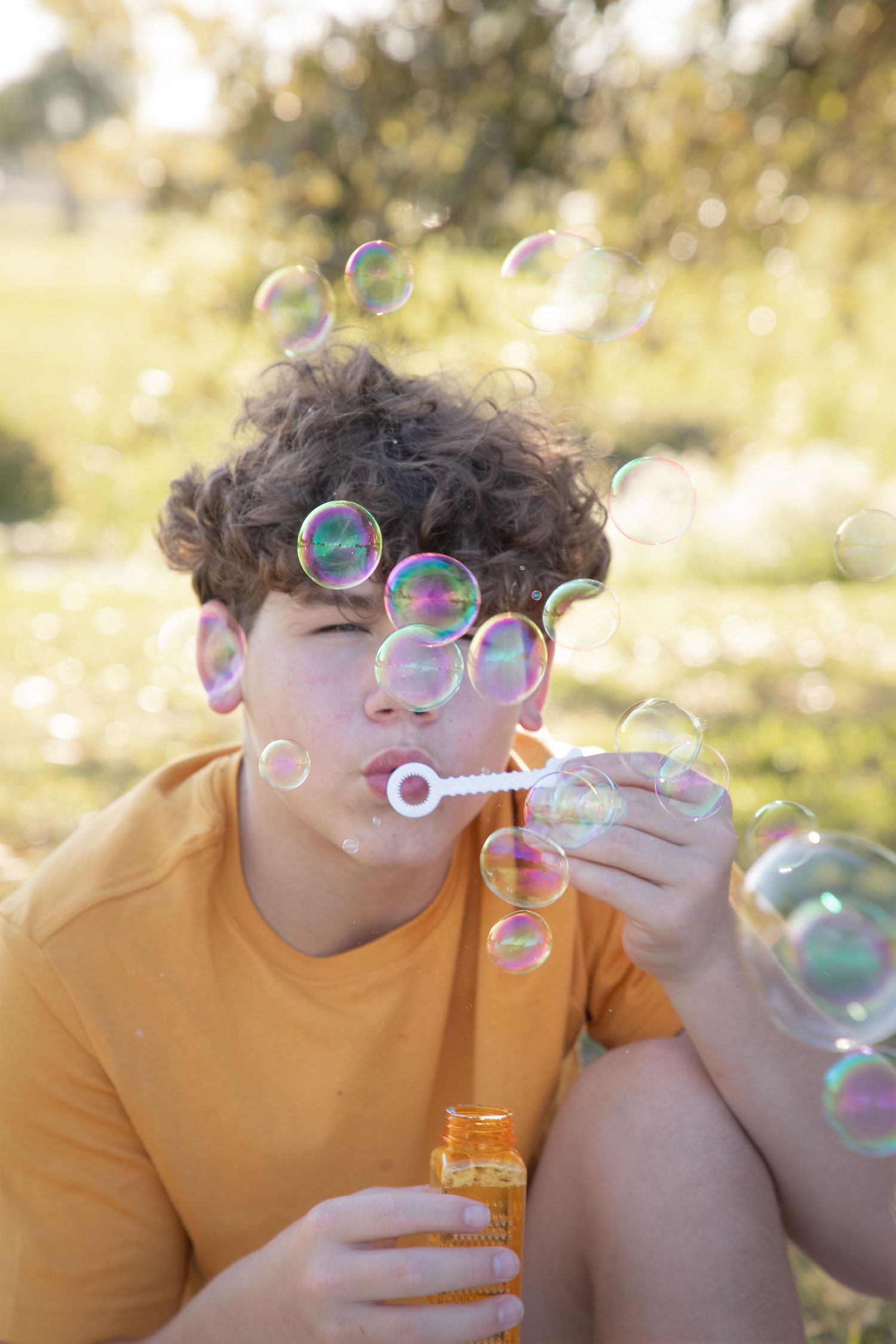 Iridescent soap bubbles float in golden sunlight as someone in an orange t-shirt blows bubbles outdoors.