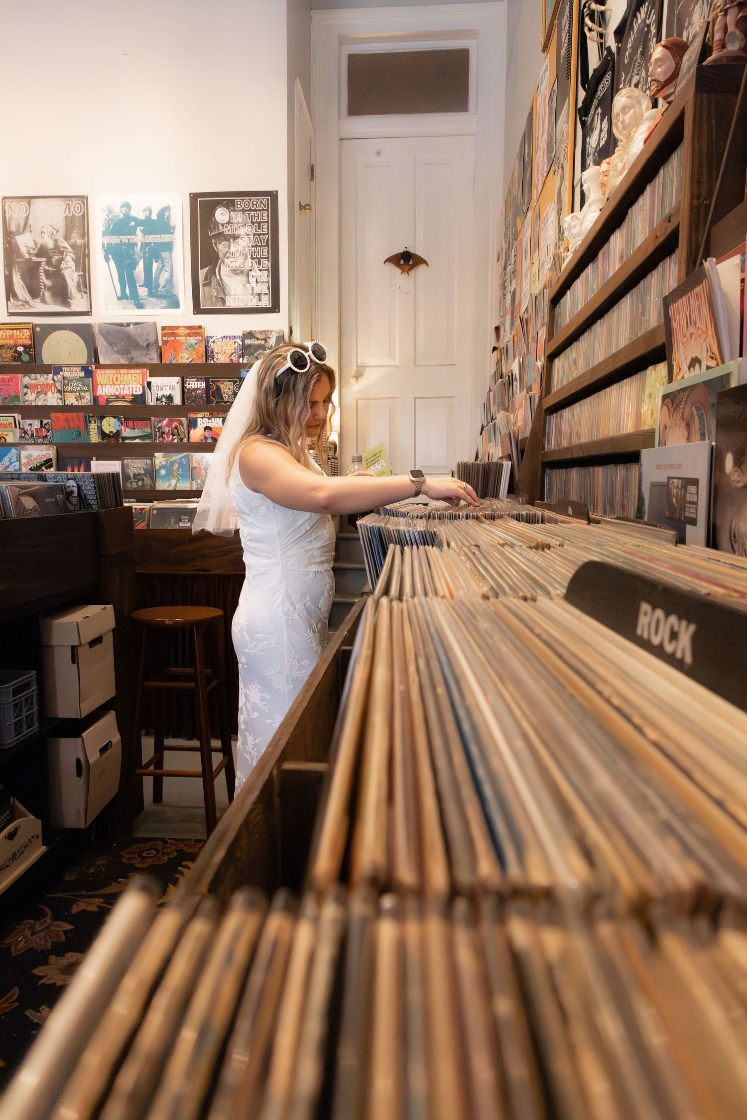 Bride in white dress browsing vinyl records in a cozy record shop with rock section visible in foreground.