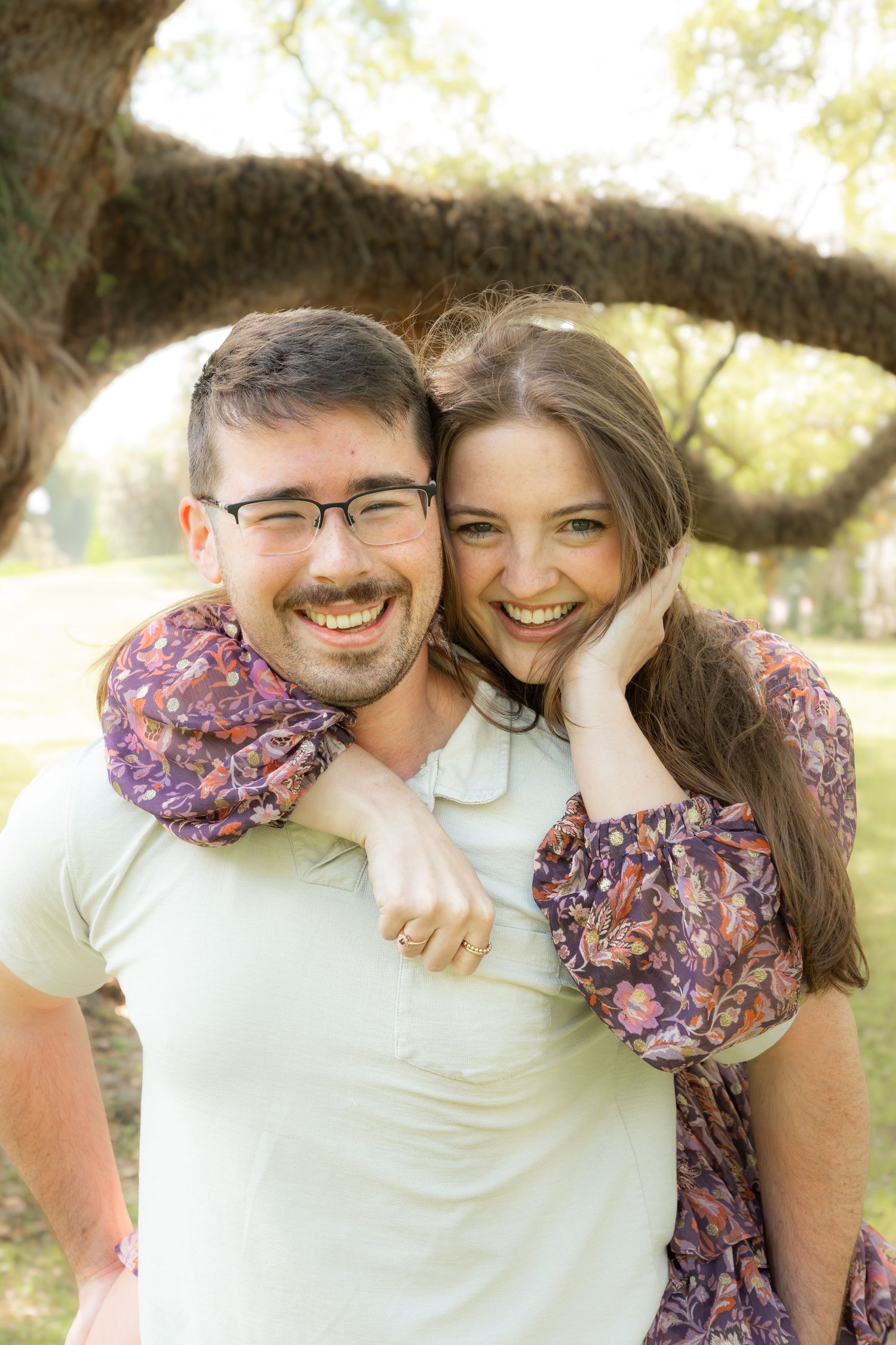 A smiling couple embraces outdoors under a large tree, woman in floral dress rests her head on man's shoulder.