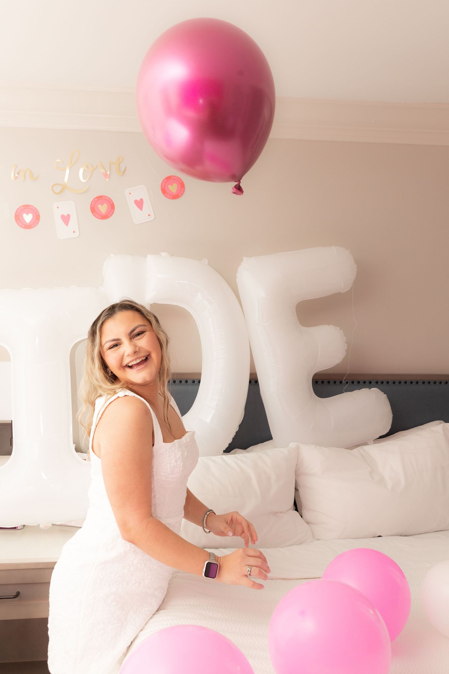 Smiling bride in white dress sitting on bed surrounded by pink balloons and white letter balloon decorations.