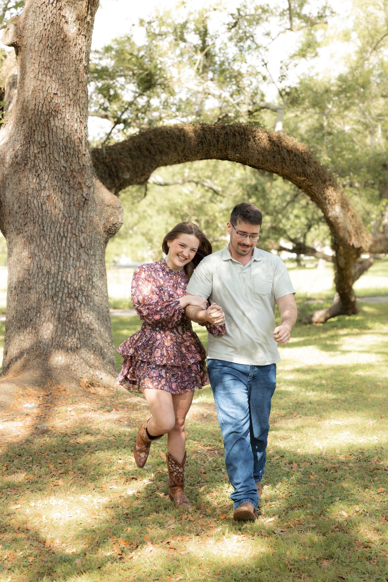 A smiling couple holds hands while walking through a sunlit park beneath a curved tree arch.