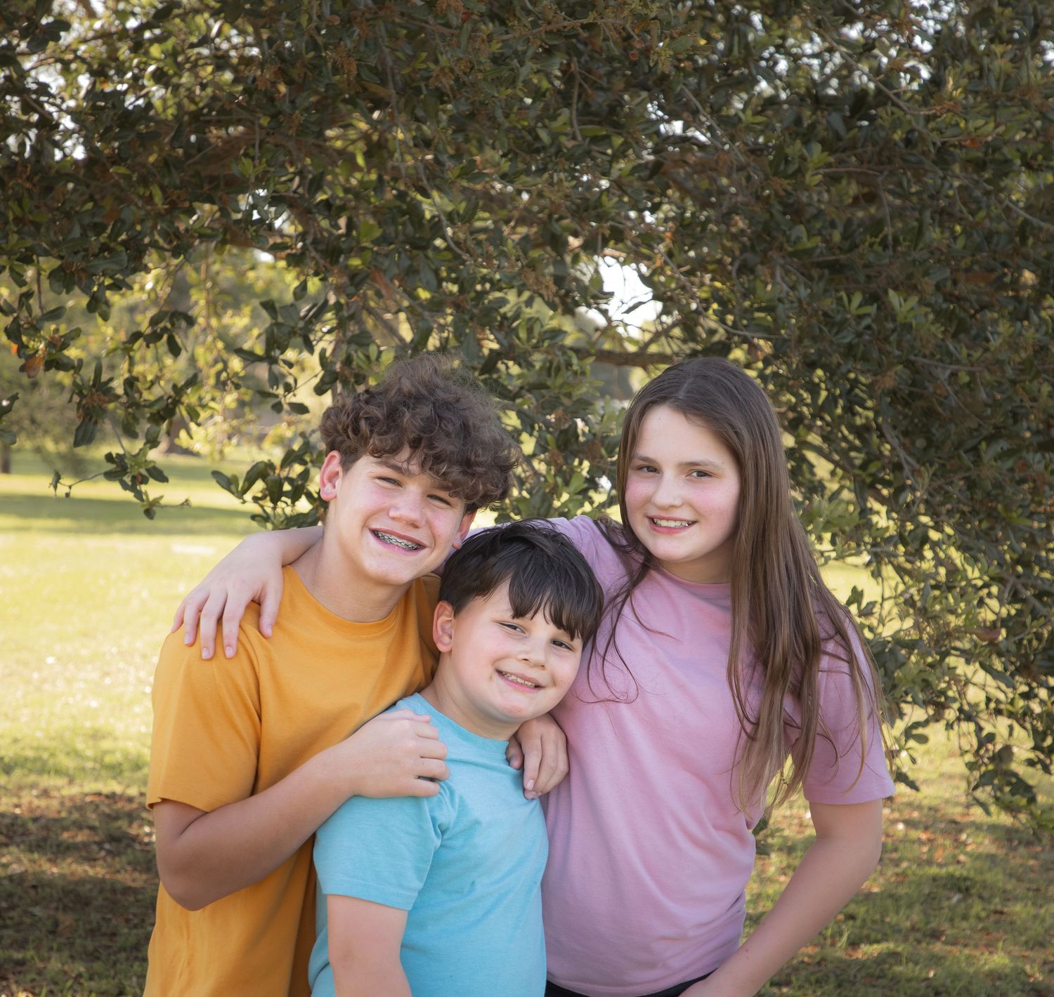 Three people share joyful moments together outdoors under tree branches on a sunny day.
