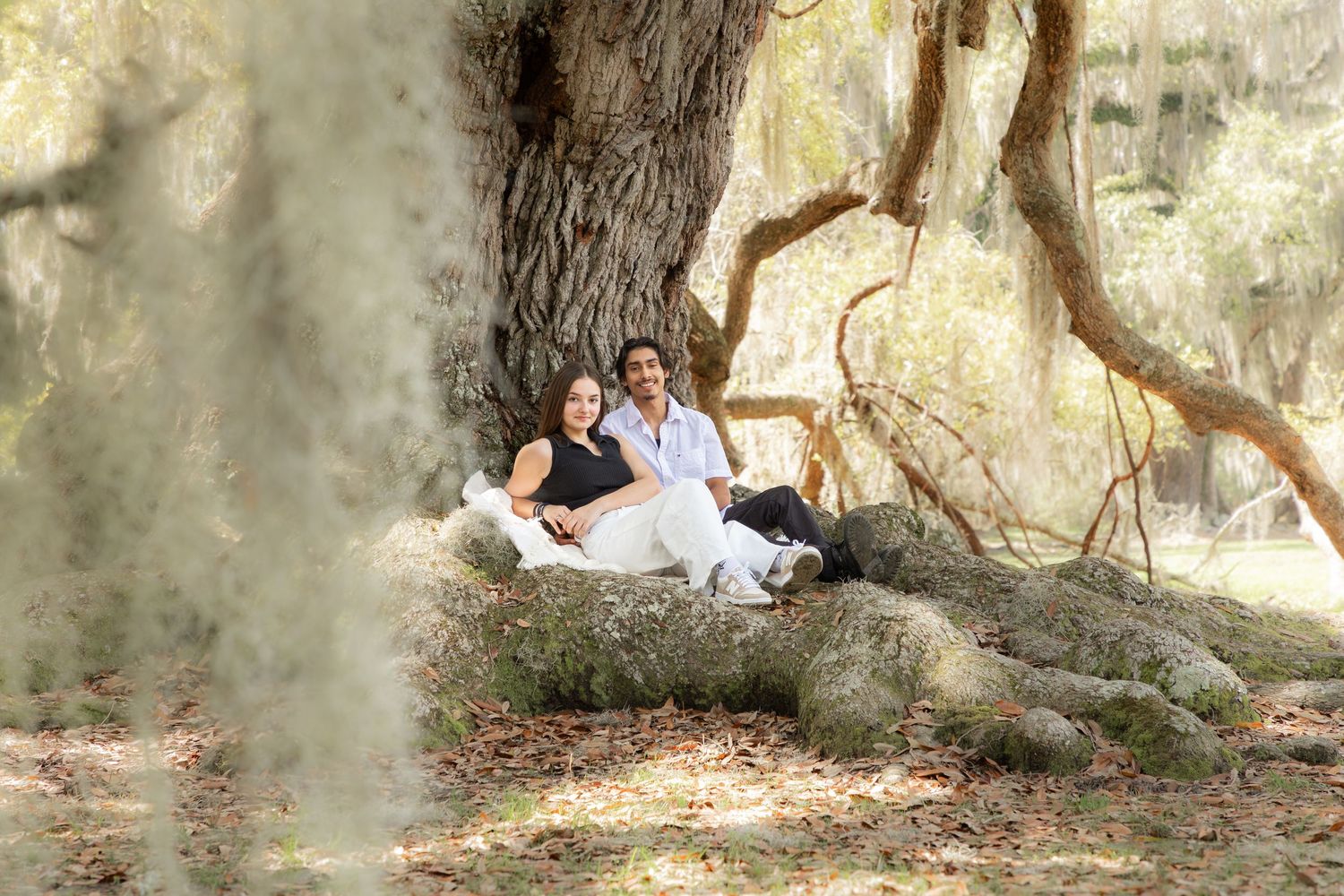 Couple sitting together against large tree roots in a scenic outdoor park with Spanish moss.