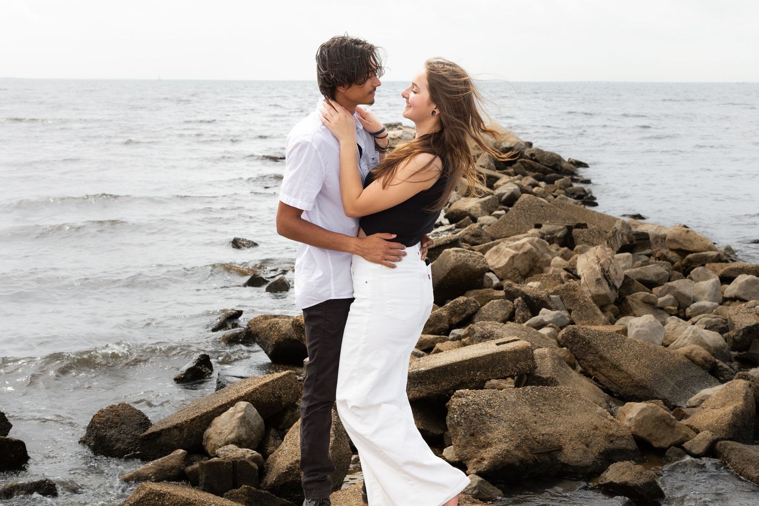 Romantic couple embracing on rocky shoreline with ocean waves in the background on a cloudy day.