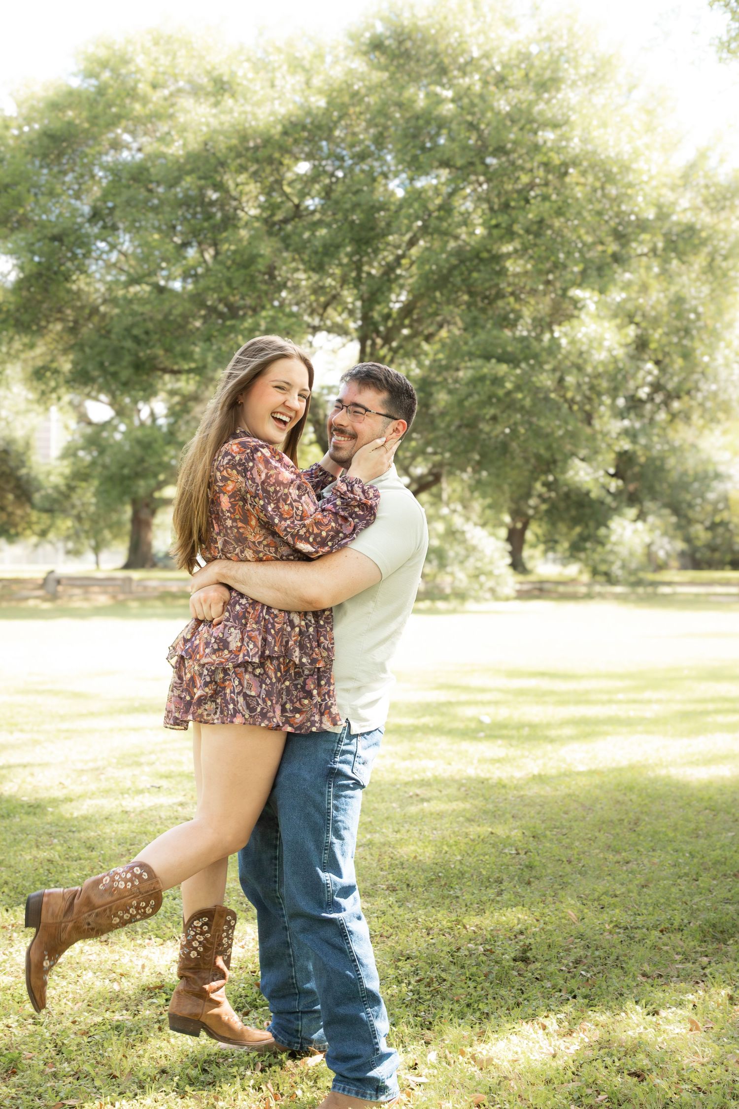 A laughing couple embraces in a sunny park, woman in floral dress and cowboy boots, man in jeans and white shirt.