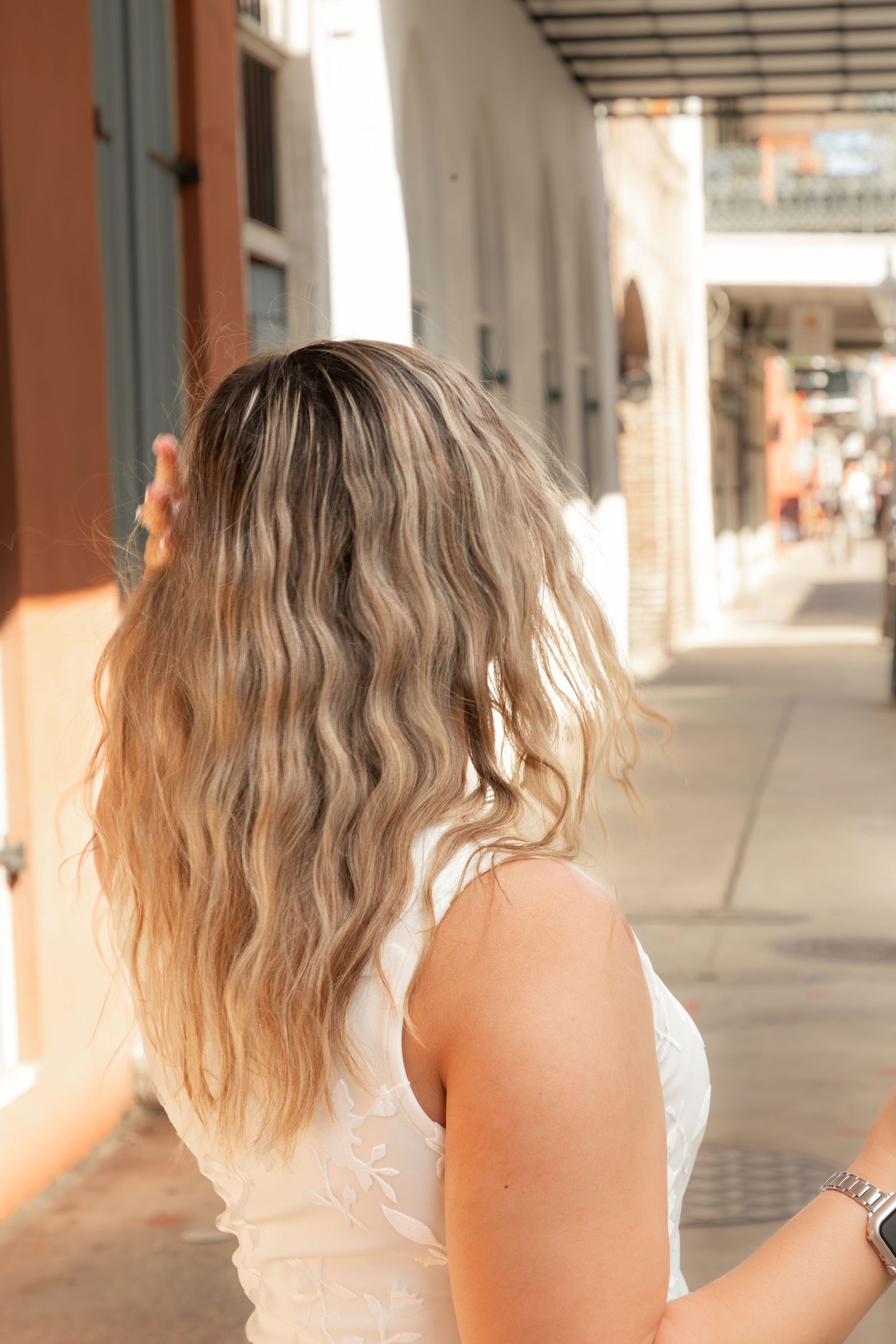 Woman with wavy blonde ombre hair wearing a white top and silver watch on a sunny urban street.