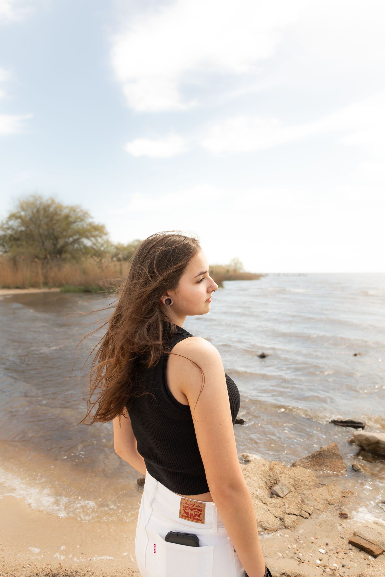 Young woman in black top and white Levis jeans standing on rocky beach shore with wind blowing her long brown hair.