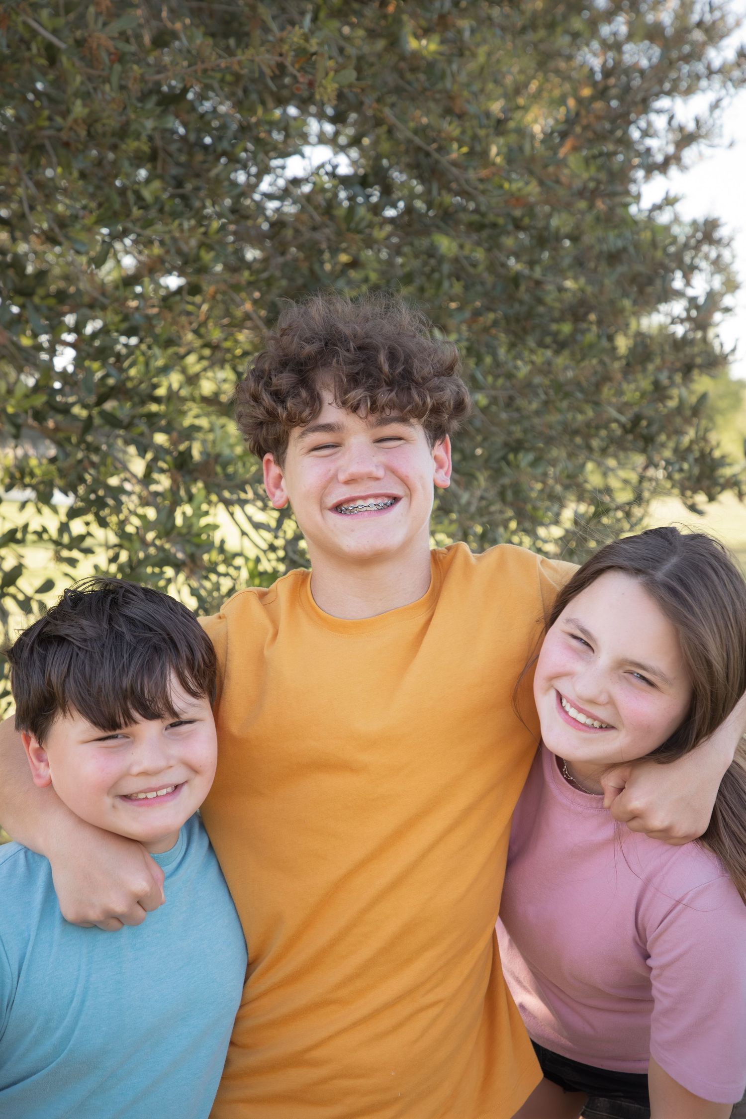 Three young people wearing colorful t-shirts smile together outdoors with trees in the background.
