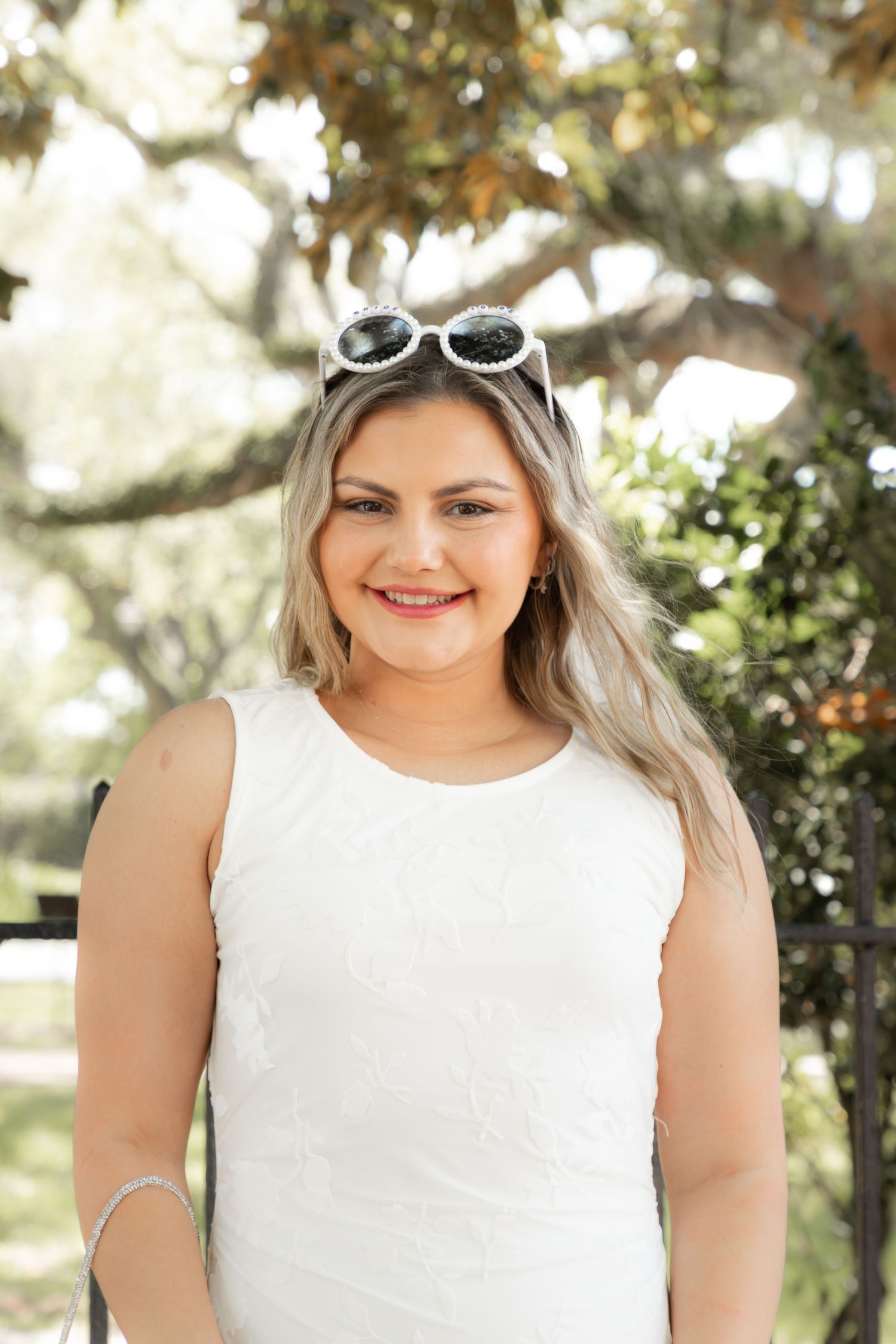 Young woman in white sleeveless dress with sunglasses on head, smiling outdoors under trees.