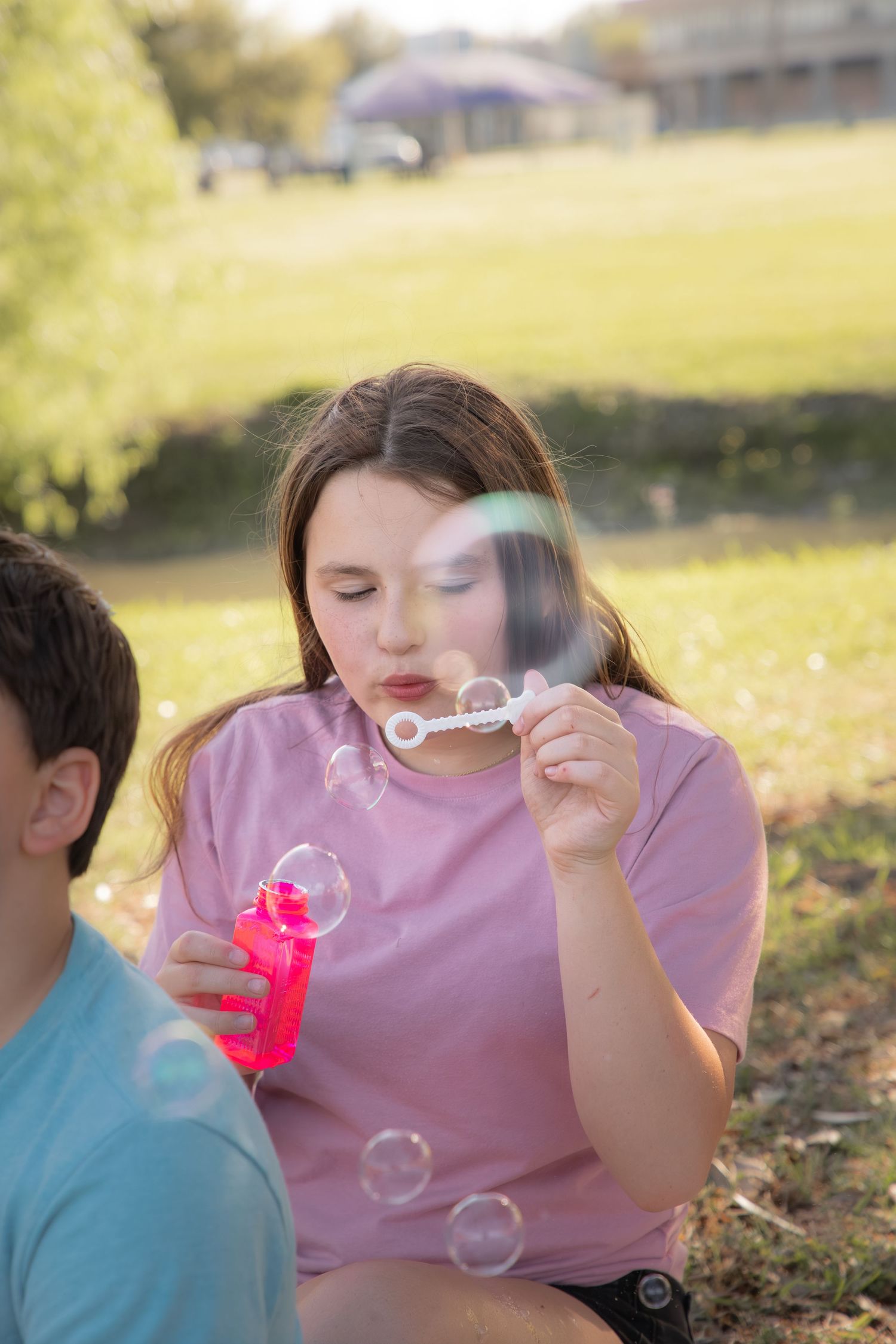 Someone in a pink t-shirt blows soap bubbles outdoors on a sunny day in a grassy park setting.
