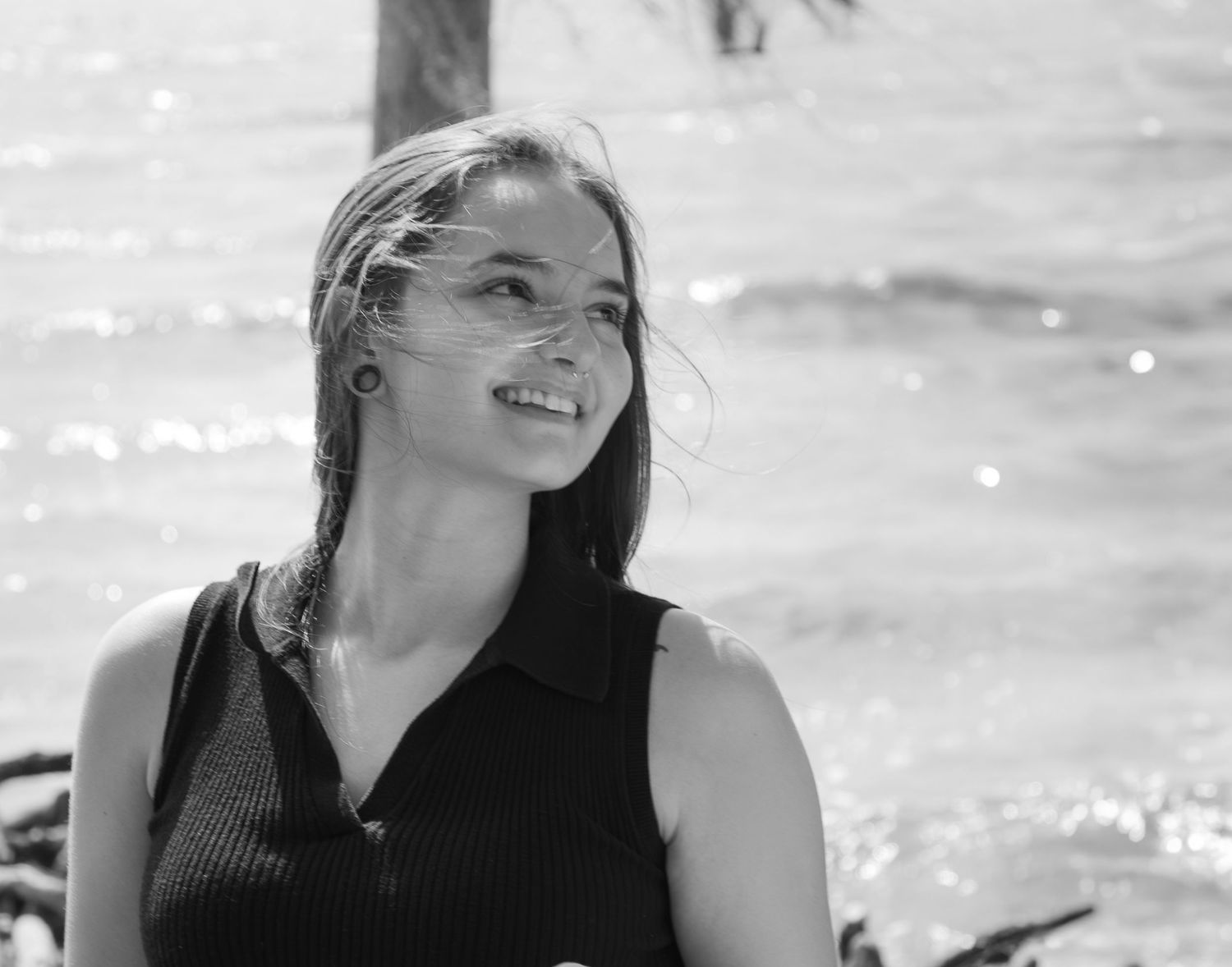 A woman in a black sleeveless top smiles while looking off to the side against a bright, sunlit background.