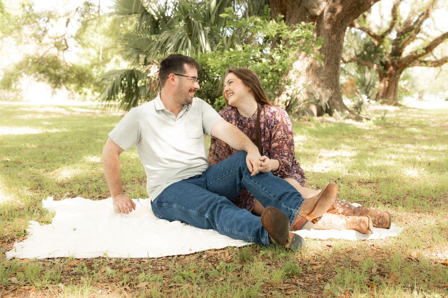 Couple sitting on white blanket in park, smiling at each other under large tree on sunny day.