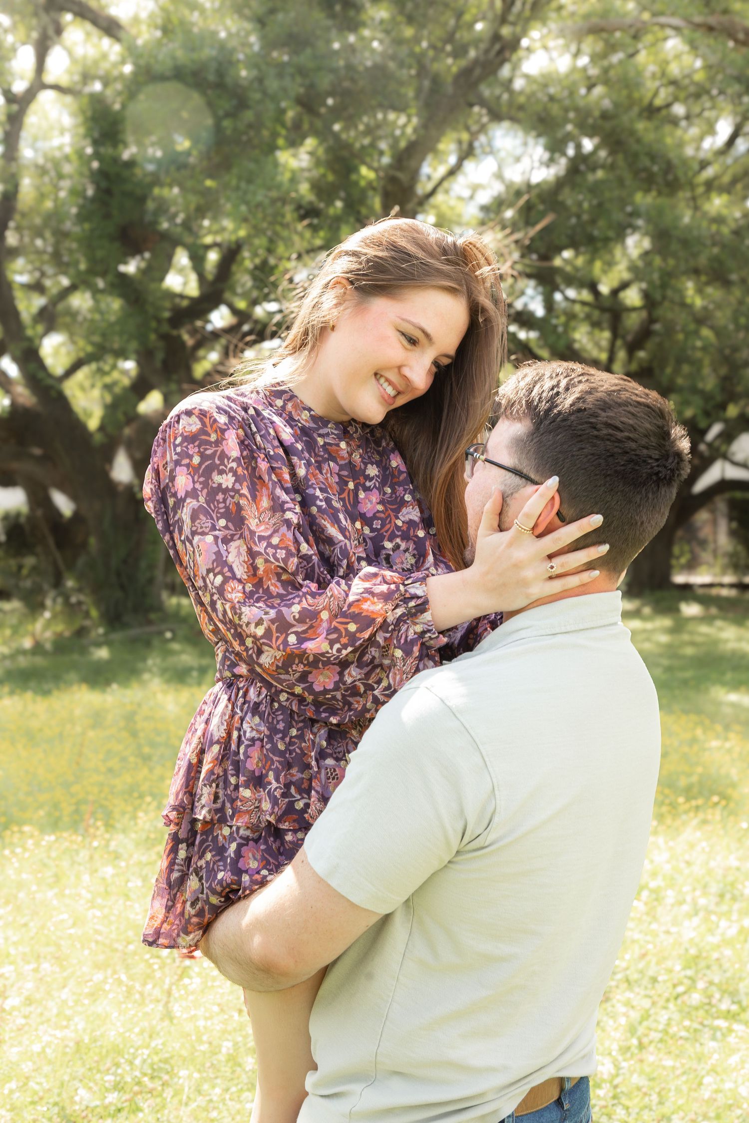 A man lifts a smiling woman in a floral dress outdoors, as she tenderly cups his face in a sunny park.