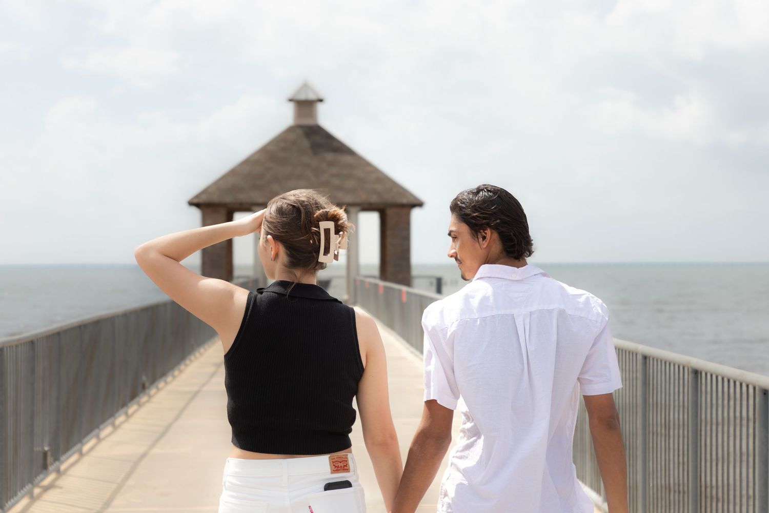Couple holding hands walking on a pier toward a gazebo overlooking the ocean on a cloudy day.