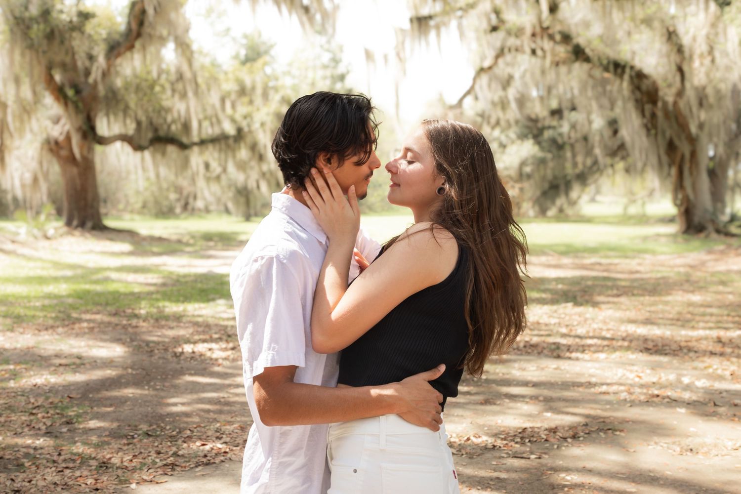 Couple sharing a romantic kiss outdoors under Spanish moss-draped trees in a sunlit park setting.