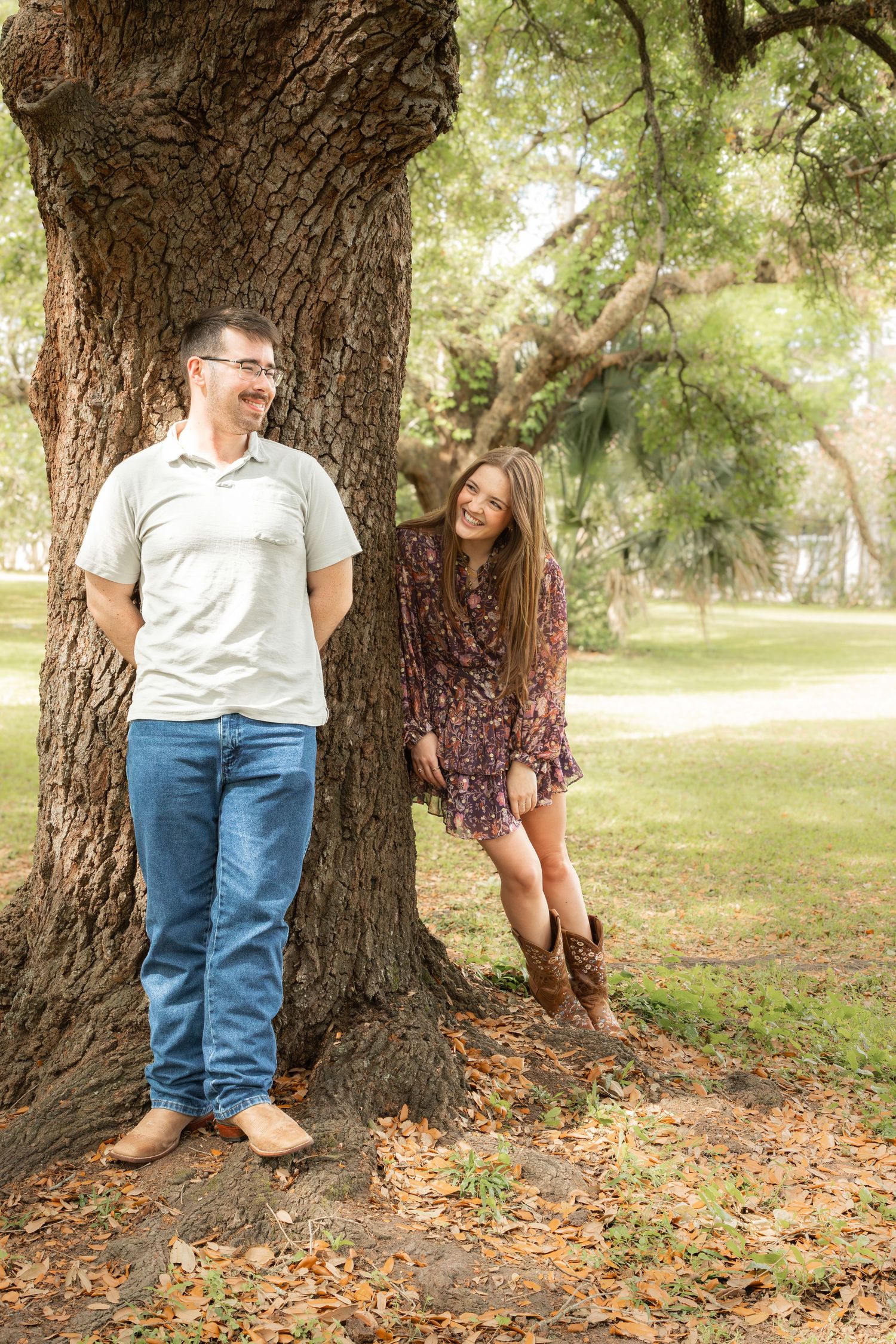 Couple smiling and leaning against a large tree in a park, woman in floral dress and cowboy boots.