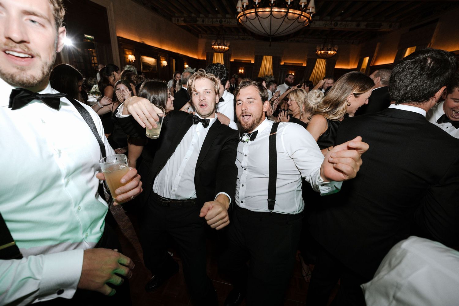 Guests in black tuxedos and suspenders celebrate energetically on the wedding dance floor.