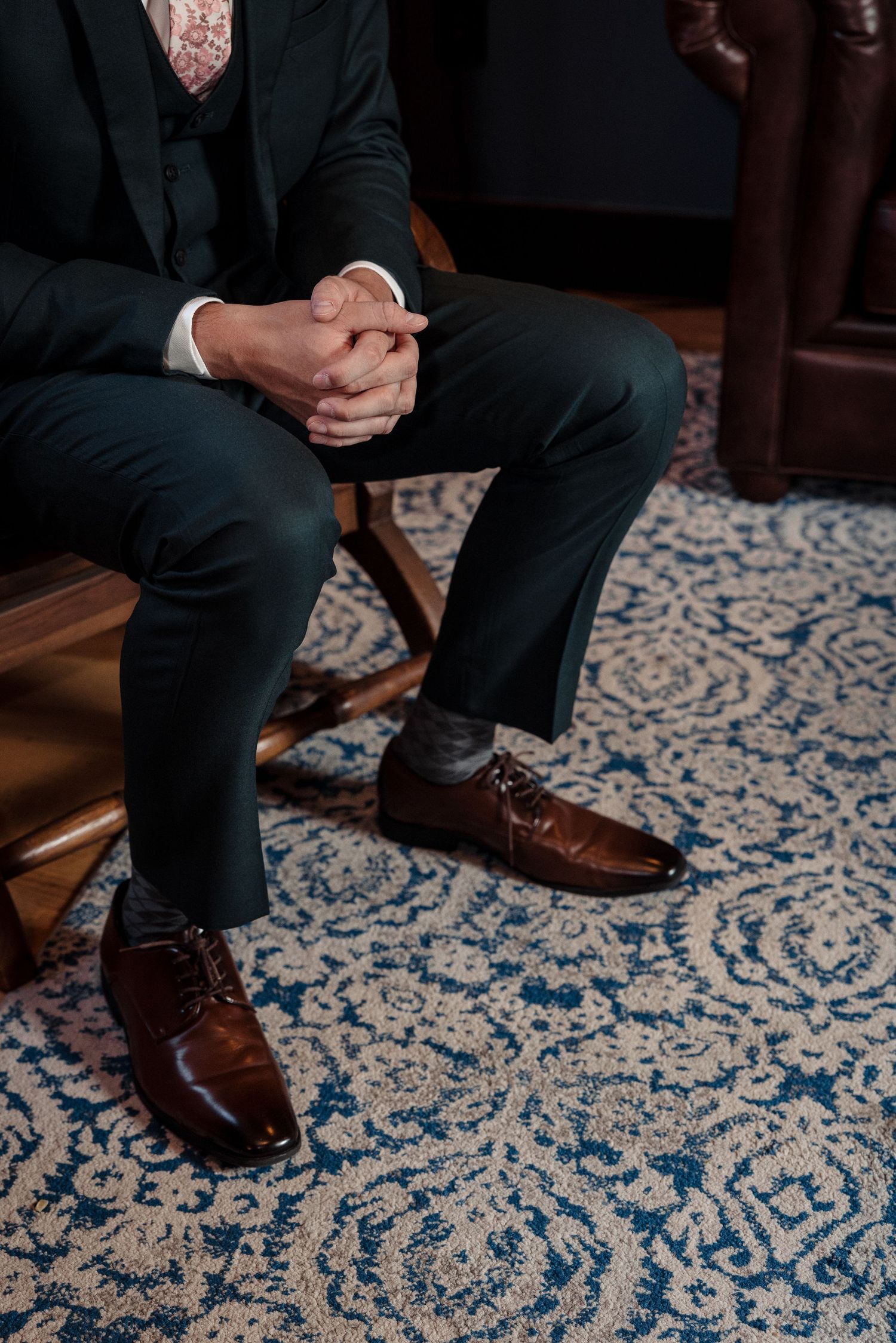 Groom in dark suit sits with clasped hands wearing polished brown leather dress shoes.