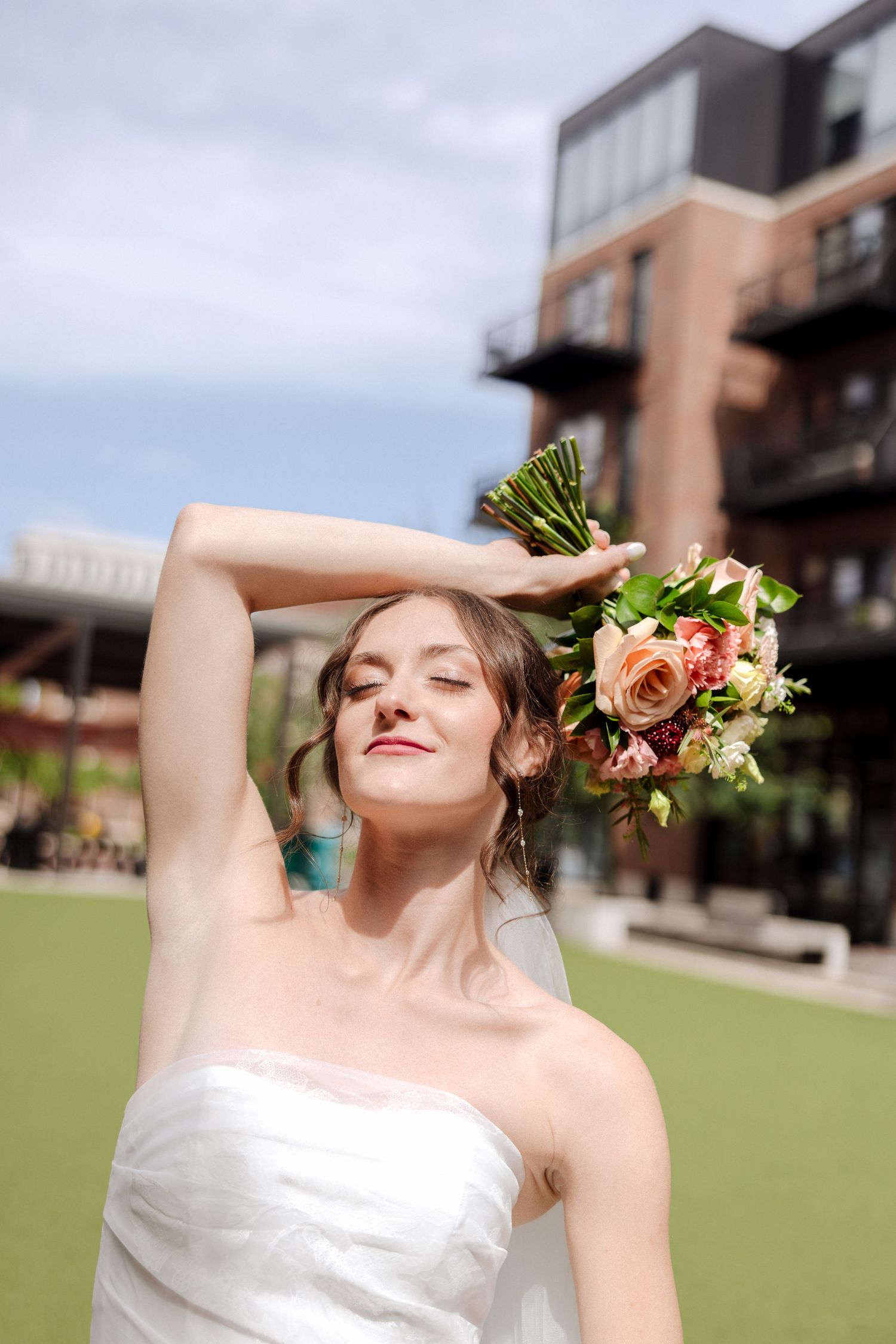 Bride in strapless white gown posing with peach rose bouquet raised near head on sunny outdoor green lawn.