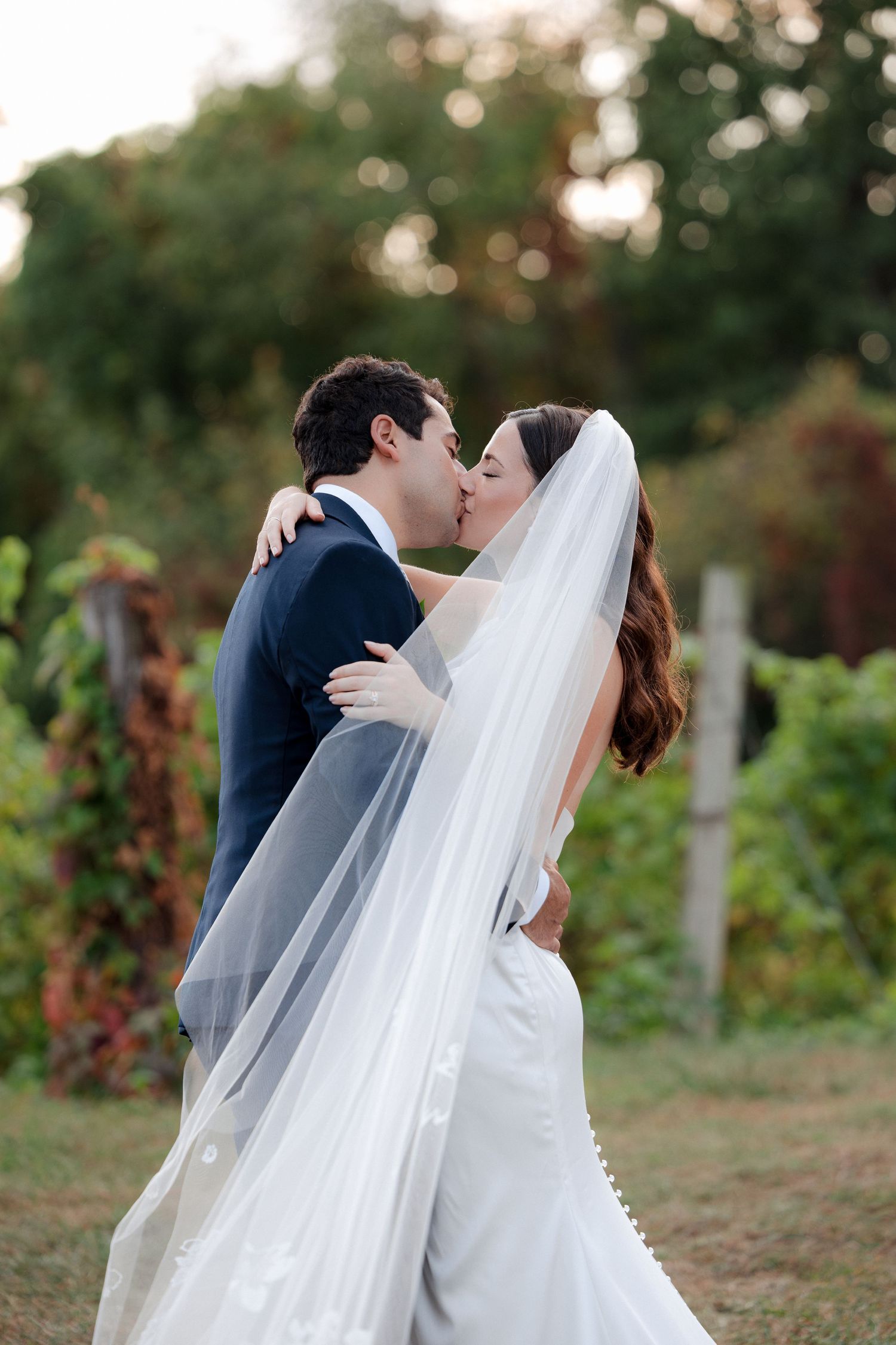 Bride and groom sharing romantic kiss in vineyard, bride wearing long veil with pearl trim.