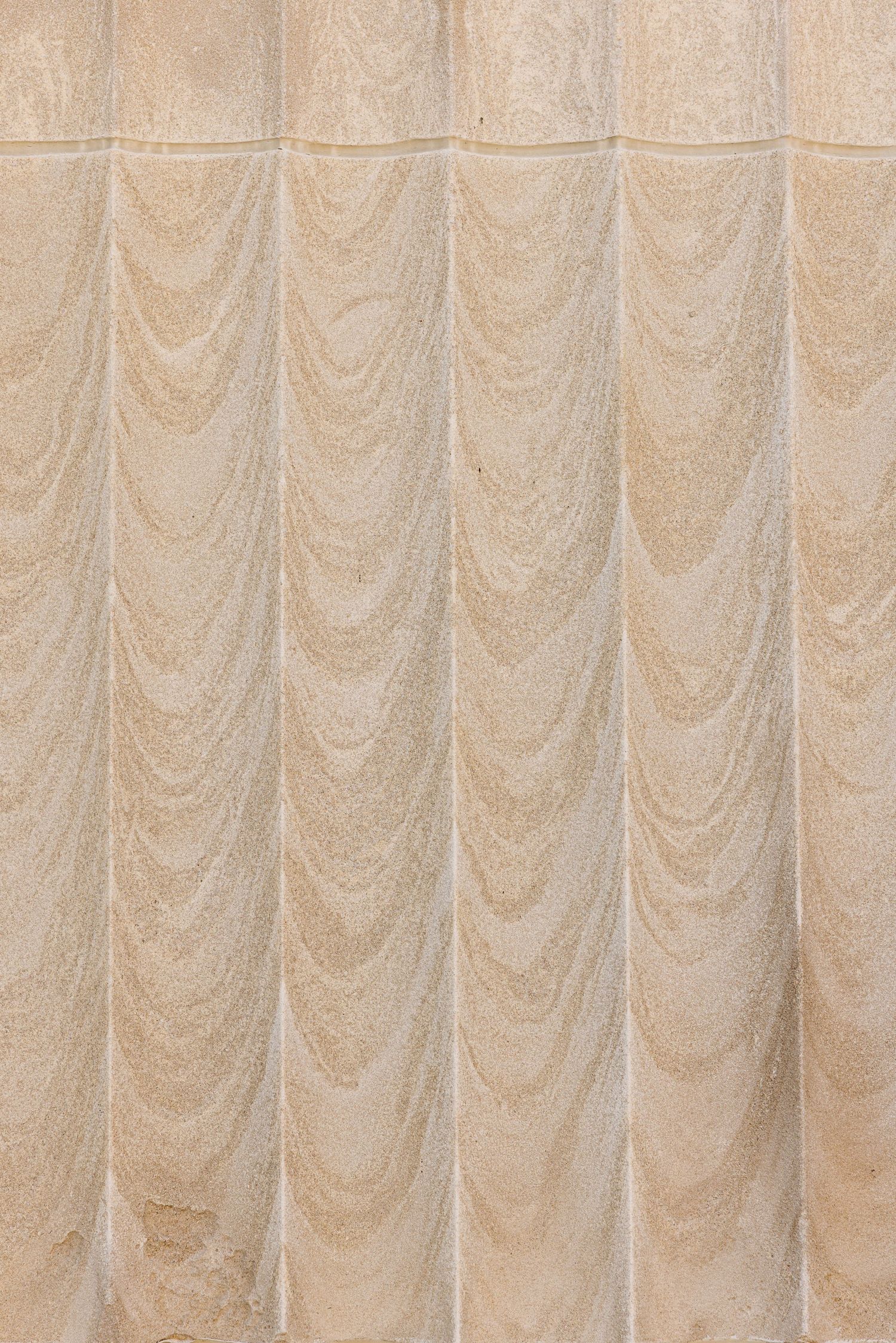 Beige stone wall with carved draped fabric relief pattern in repeating swag design, classical architectural detail texture.