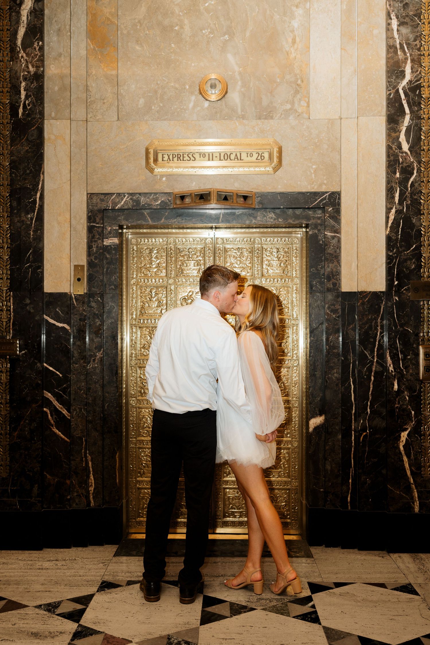 Couple sharing a romantic kiss in front of ornate gold elevator doors in a marble lobby.