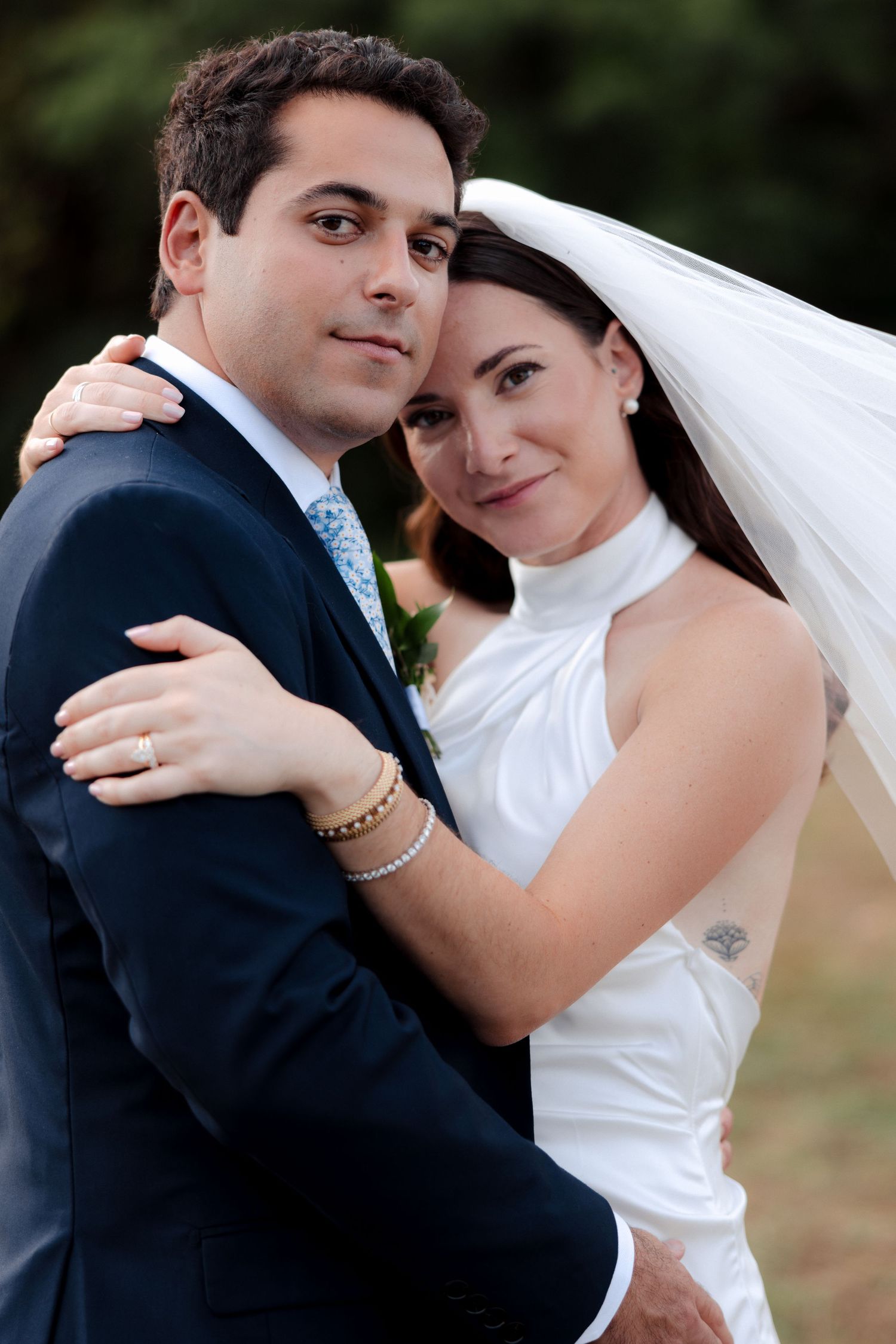 Bride in white halter gown and veil embraces groom in navy suit outdoors in a romantic wedding portrait.