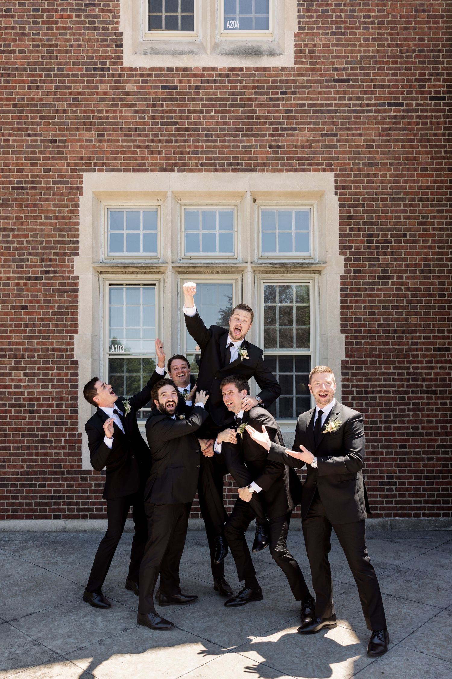 Excited groom lifted by groomsmen in black tuxedos outside a brick building at a wedding.