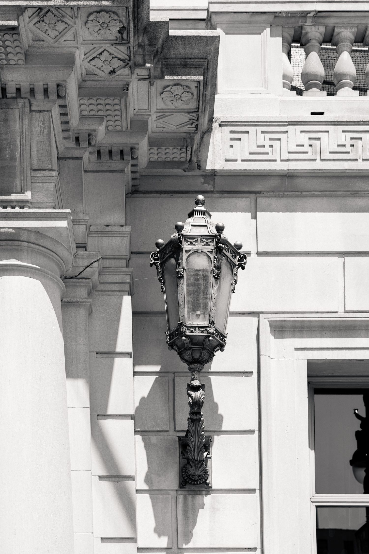 Black and white close-up of an ornate vintage street lamp mounted on a classical stone building facade.