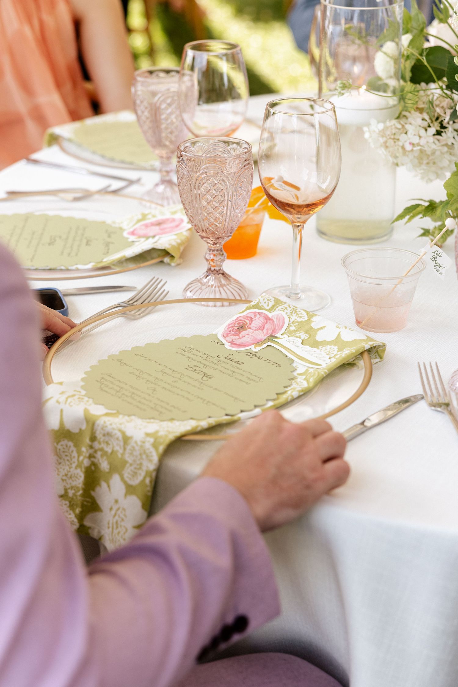 Elegant outdoor wedding reception table setting with pink glassware, floral menus, and green napkins on white tablecloth.