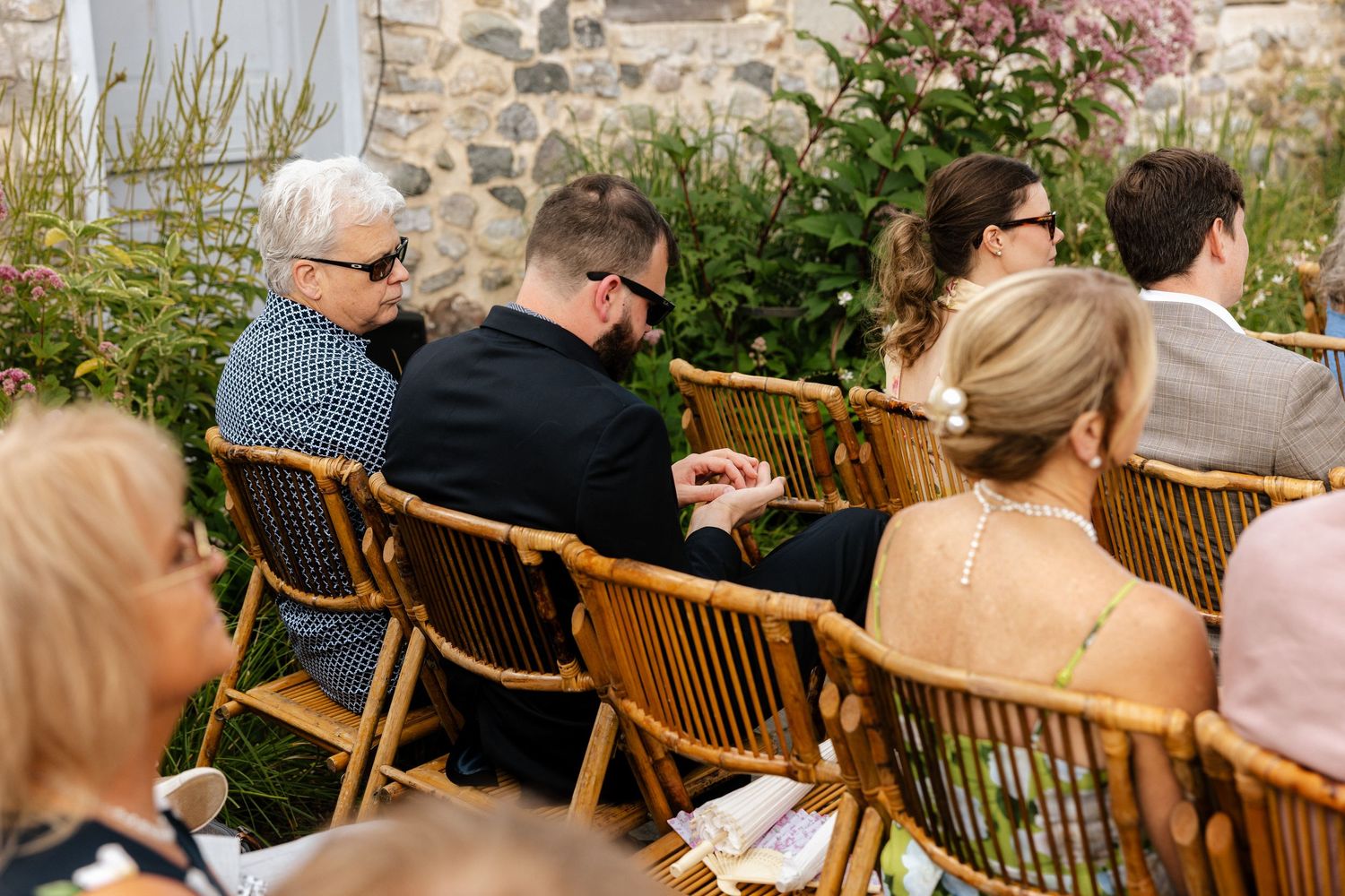 Guests seated in rattan chairs at an outdoor wedding ceremony beside a stone wall with lush greenery.