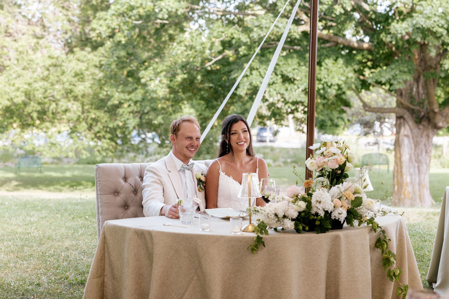 Bride and groom smiling at sweetheart table with white floral arrangements at outdoor reception.