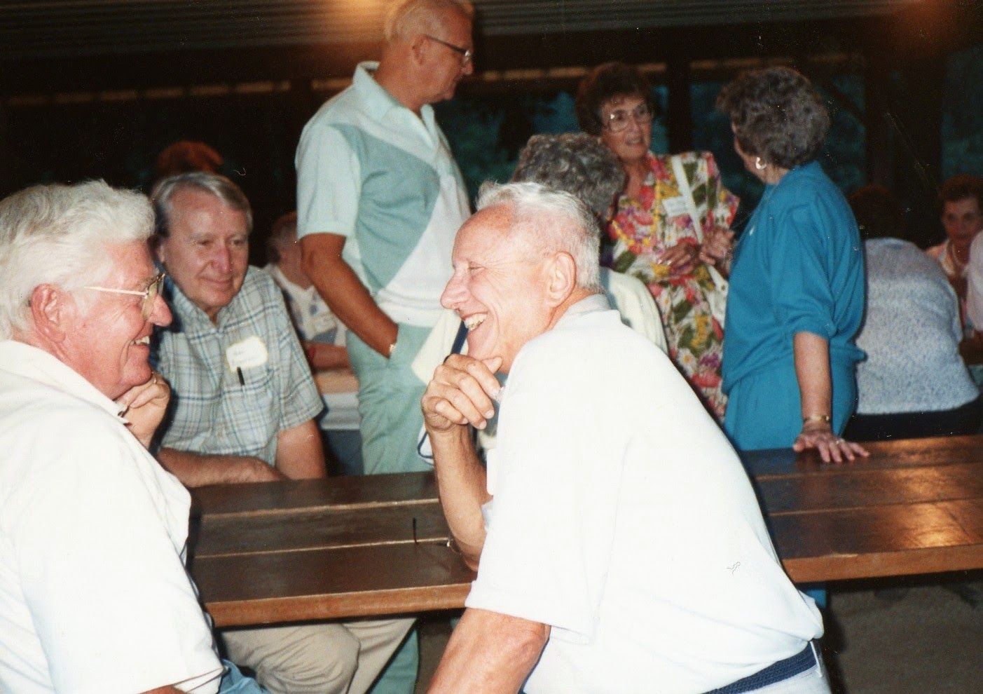 Elderly guests laugh and converse around wooden picnic tables at a casual indoor social gathering.