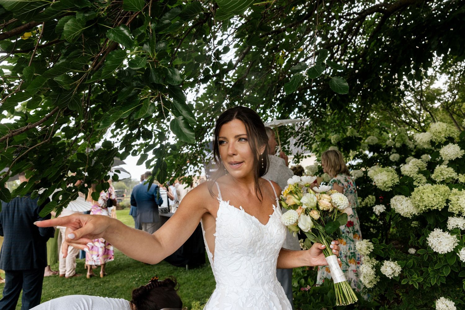 Bride in white lace gown holds bouquet outdoors under lush green trees at elegant garden wedding.
