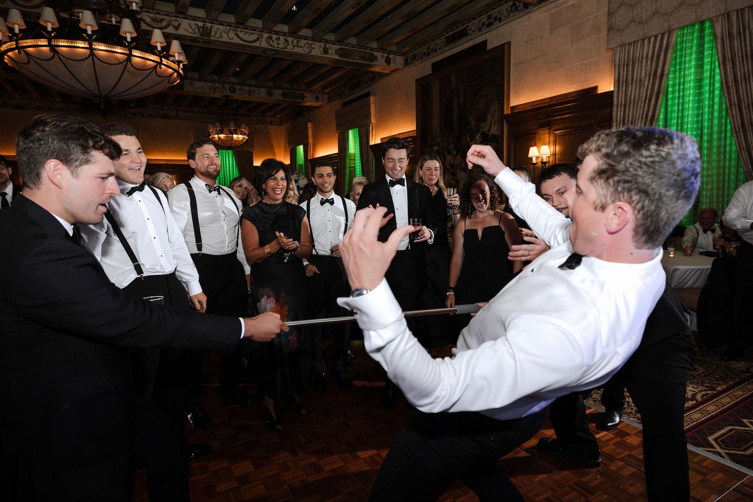 Wedding guests in tuxedos cheer as a man performs a limbo dance at the reception.