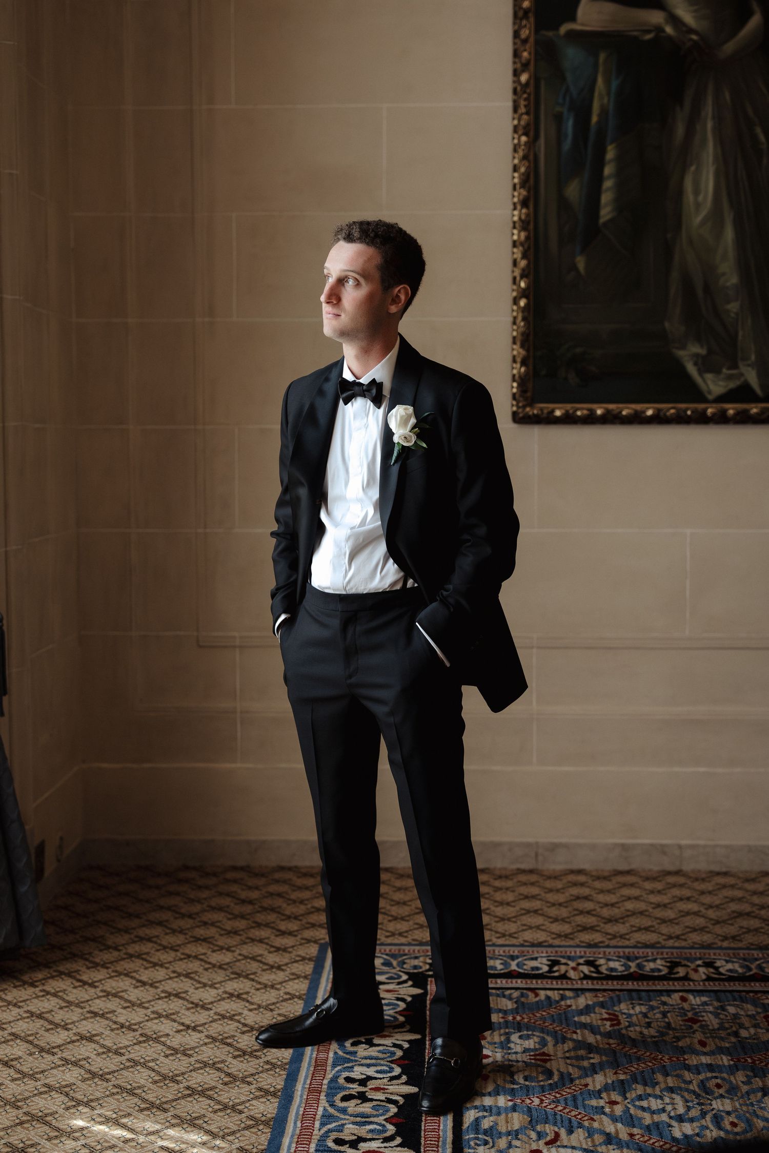 Groom in black tuxedo with white boutonniere stands in elegant hall.