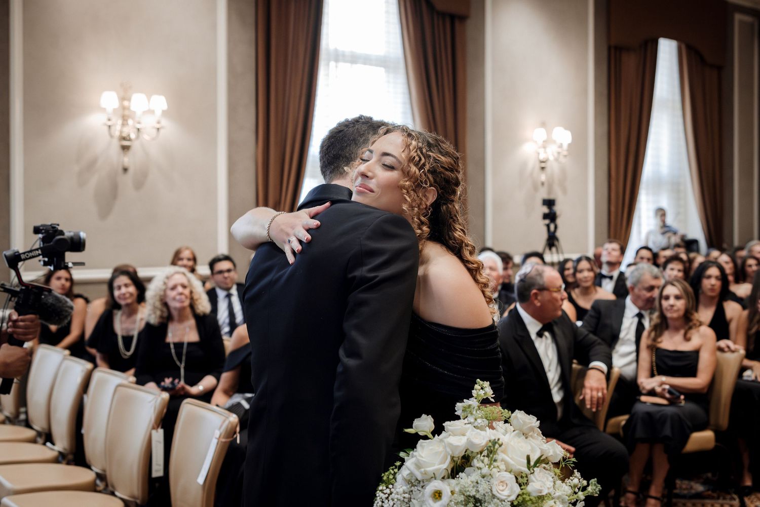 Emotional embrace between two guests in formal attire during an elegant hotel ballroom wedding ceremony.